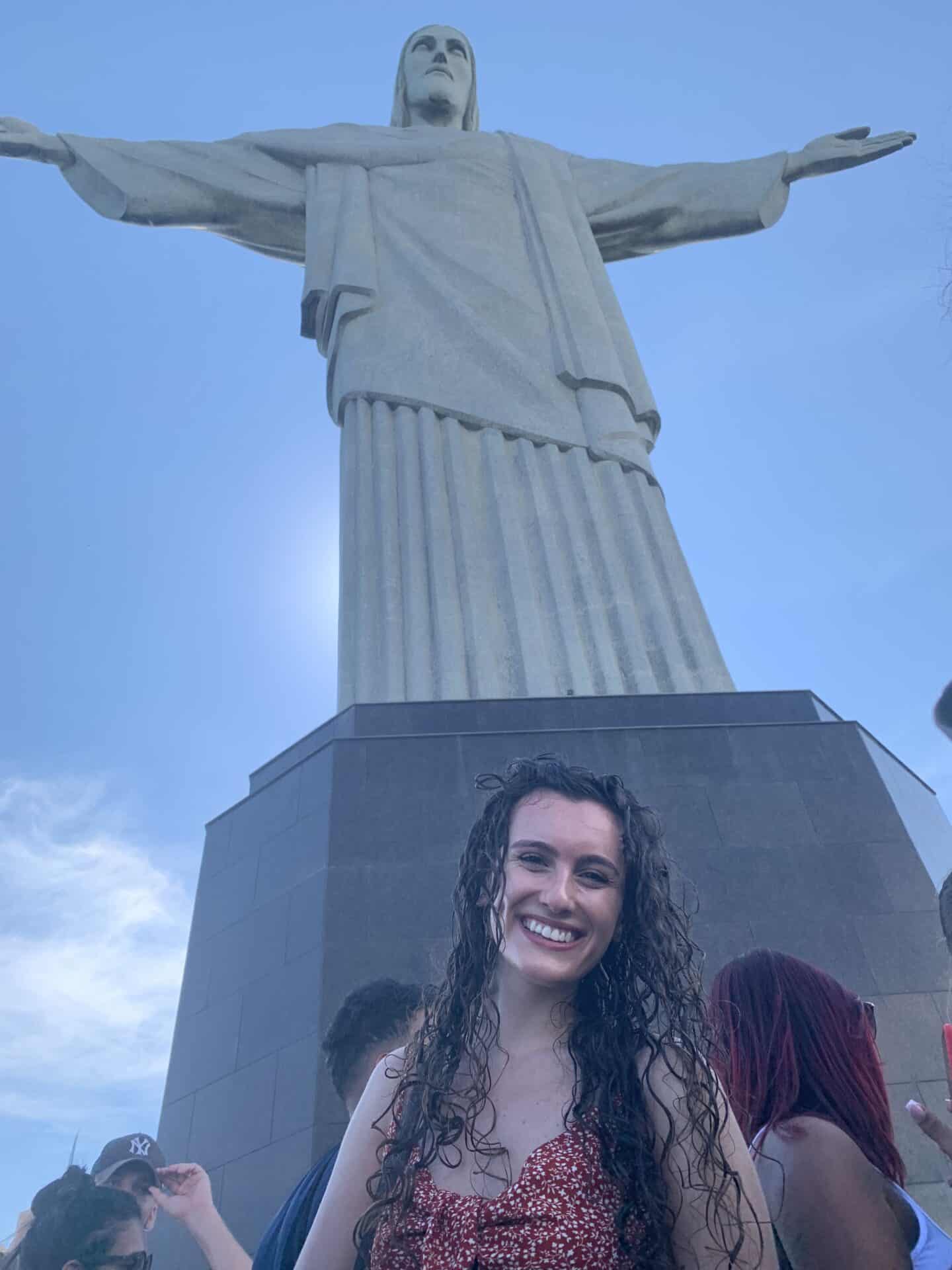 Tess, wearing a red shirt, smiles in front of the Christ the Redeemer statue in Rio de Janeiro, Brazil.