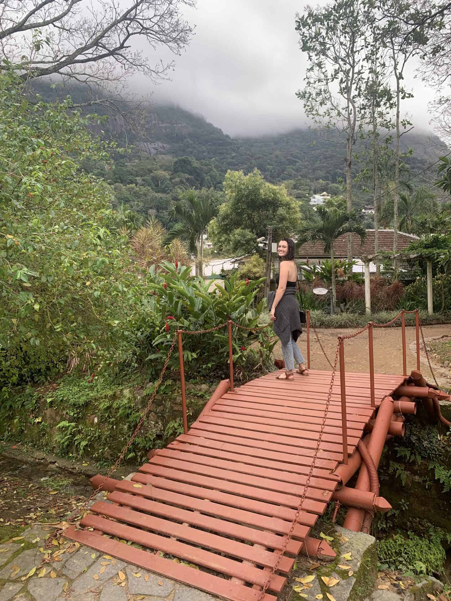 A woman stands on a red bridge in the Botanical Garden in Rio de Janeiro with trees and clouds surrounding her