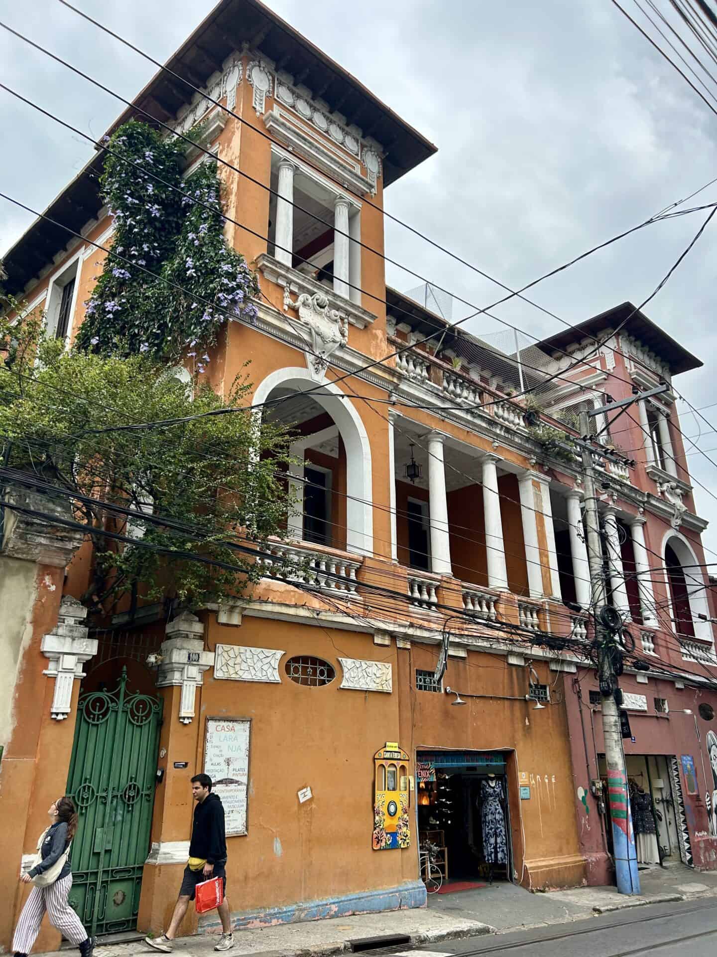 People walk past orange, colonial-style building with white columns and purple flowers in Santa Teresa, Rio de Janeiro.
