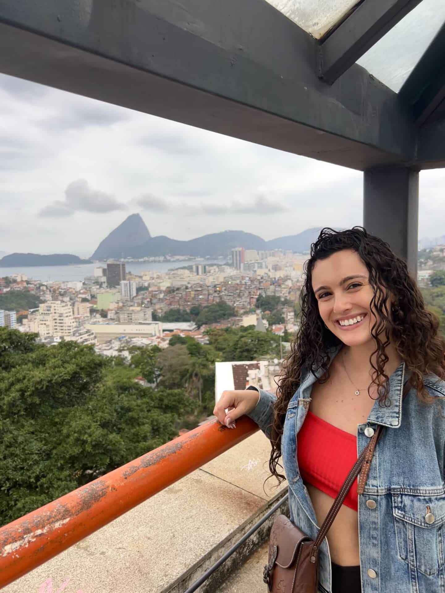 Tess stands in Parque das Ruinas in Santa Teresa, Rio de Janeiro. Behind her, is a view of the mountains and ocean on a cloudy day.