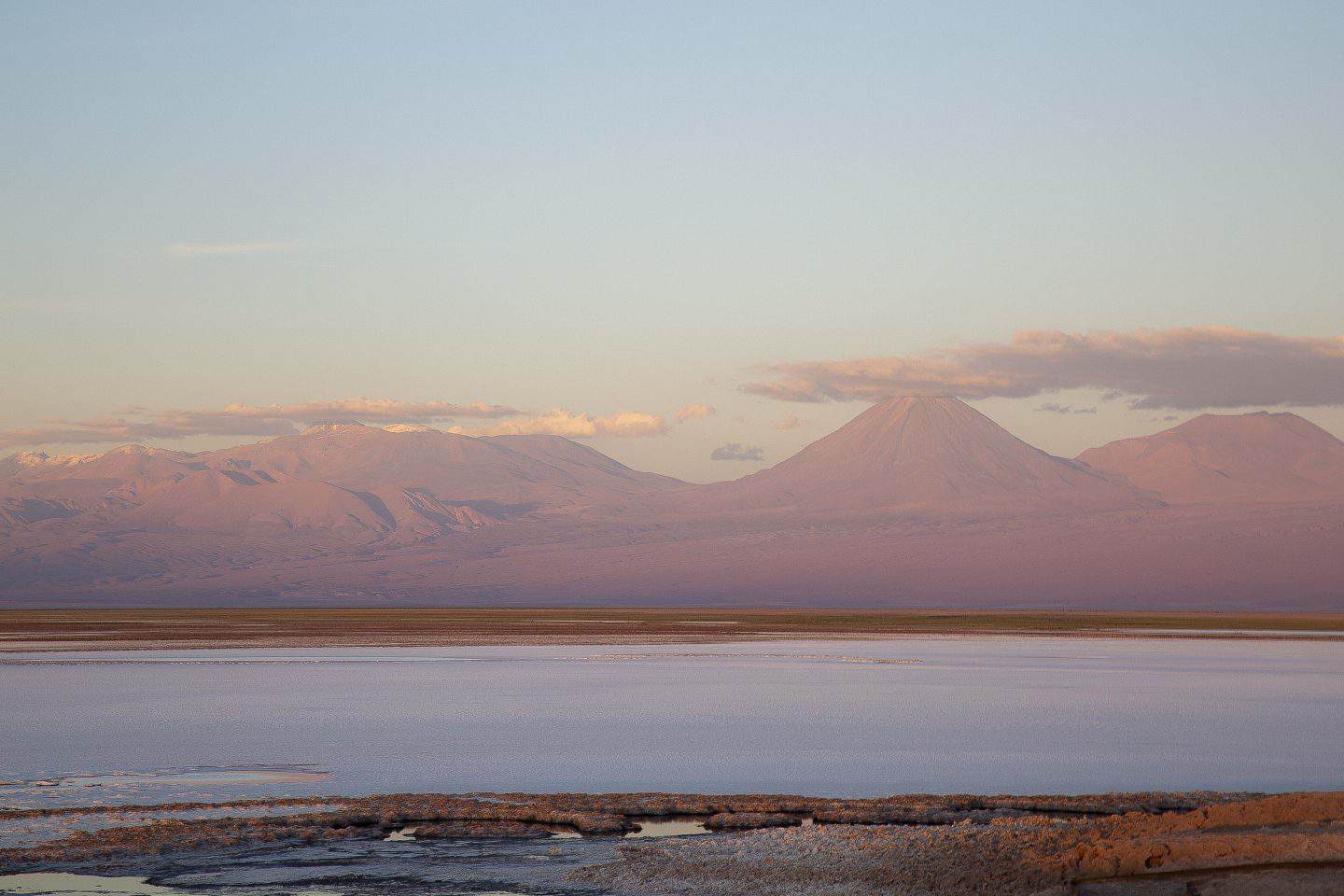A pink desert and water in the Atacama Desert in Chile with pink volcanoes in the background, and two people standing on the sand.