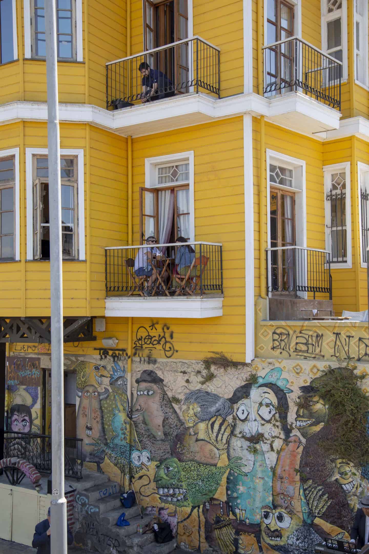A yellow building with street art at the bottom portion of it in Valparaiso, Chile. People are sitting on the balcony.
