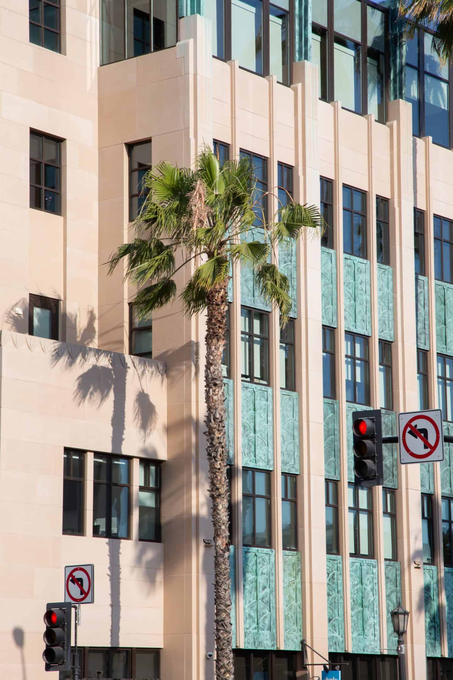 A palm tree is in front of a pink and green Art Deco building in Pasadena, California. The shadow of the tree is also visible. The building takes up the entire photo, with a couple street lights also shown.