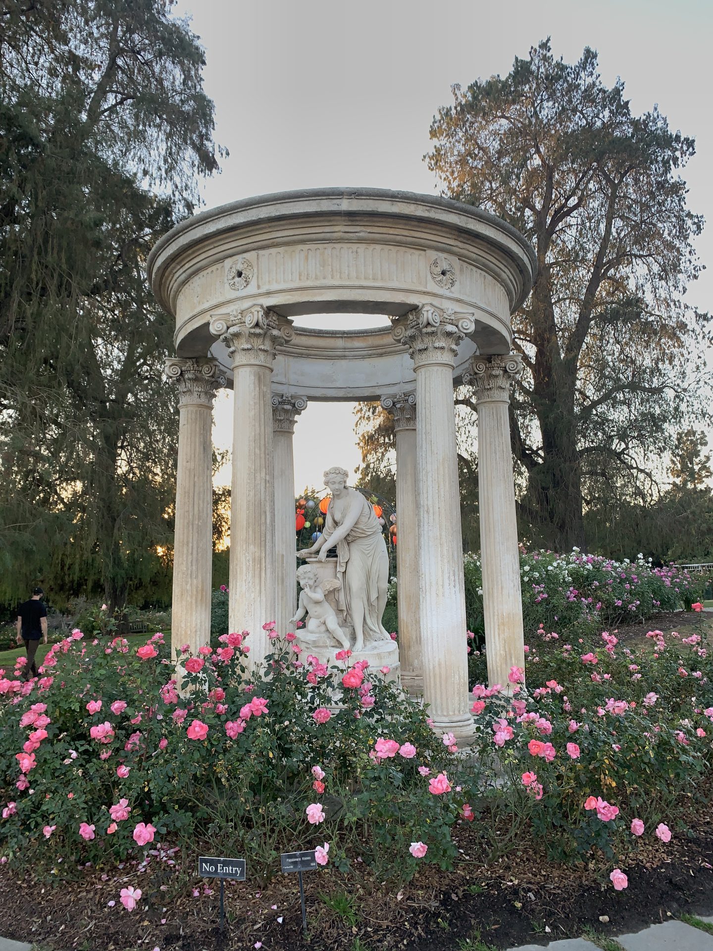 A Greek-style sculpture of a woman and a boy. Columns surround them in a circle. Pink roses are on the ground in Huntington Botanical Gardens, one of the best places to visit around Pasadena.