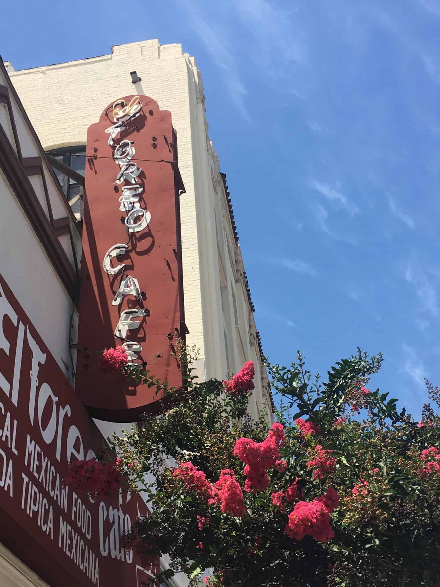 In Pasadena, California, a tree with pink flowers is at the bottom right, while a white building with red signs and white lettering are on the left, One sign is partially cut off, but "Mexican Food" and "Tipica Mexicana" are visible, while another sign says "El Toreo Cafe."