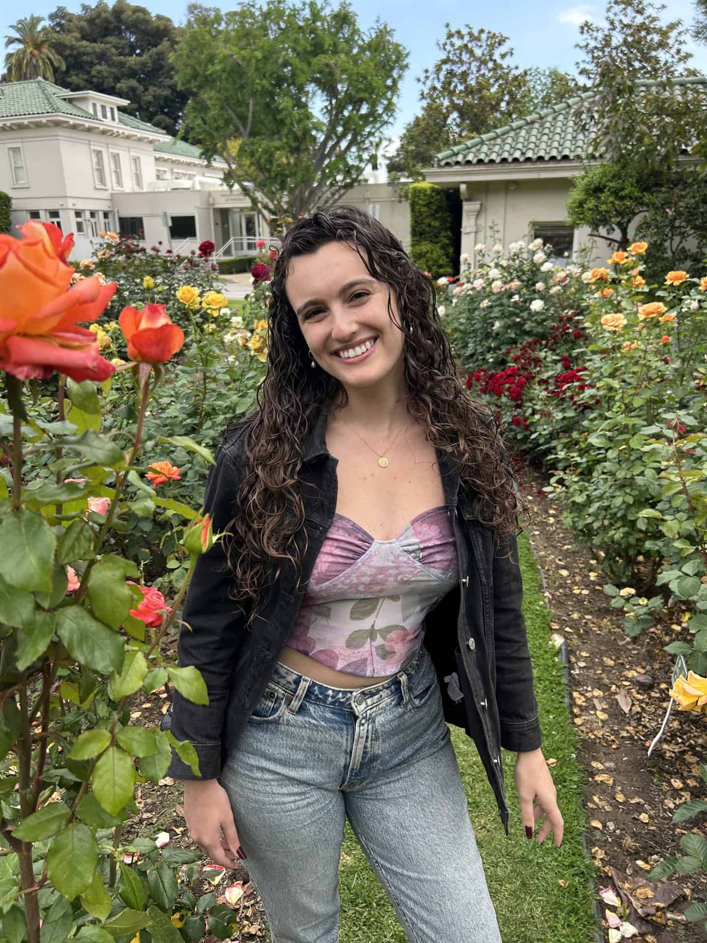 A girl wearing a flower-printed shirt, a black denim jacket, and faded blue jeans, stands in Wrigley Garden in Pasadena, California, surrounded by roses, with white buildings with green roofs in the background.