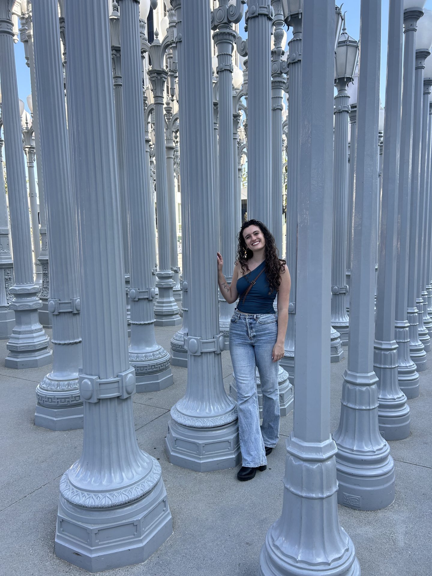 A woman wearing a one-shoulder blue top and blue flared jeans stands at the street lights display in front of the LACMA, one of the most popular tourist destinations in Los Angeles.