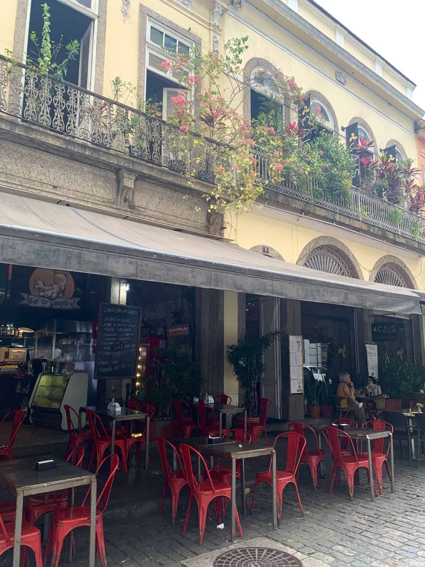A restaurant with tables and red chairs is on the bottom floor of a yellow building. The restaurant is open, Flowers line the balcony on the upper floor, and all the windows are open in Centro, Rio de Janeiro.