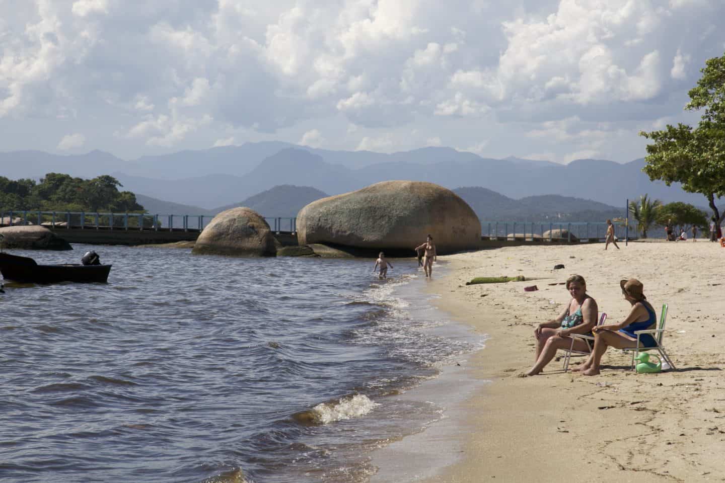 Two women sit in beach chairs near the water in Ilha de Paquetá, Rio de Janeiro. Large boulders are in the water, and layers of blue mountains are behind the water. A few other people are on the beach as well.