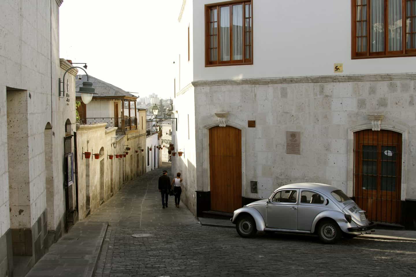 Two people walk down a cobblestone street with white buildings and a silver car parked in Arequipa, Peru