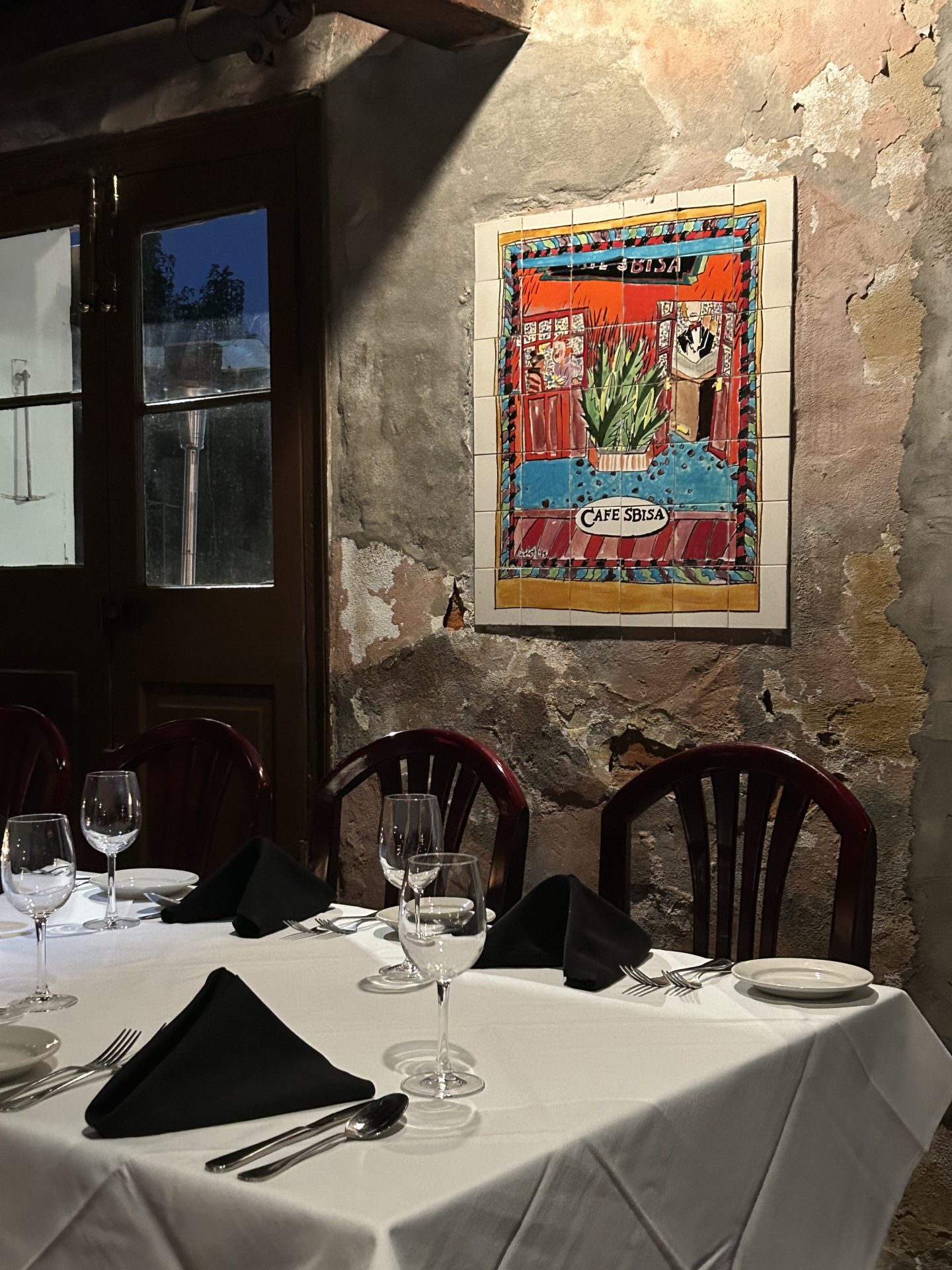A table with folded napkins and empty wine glasses, with a stone wall with artwork behind it that says "Cafe Sbisa" in New Orleans.