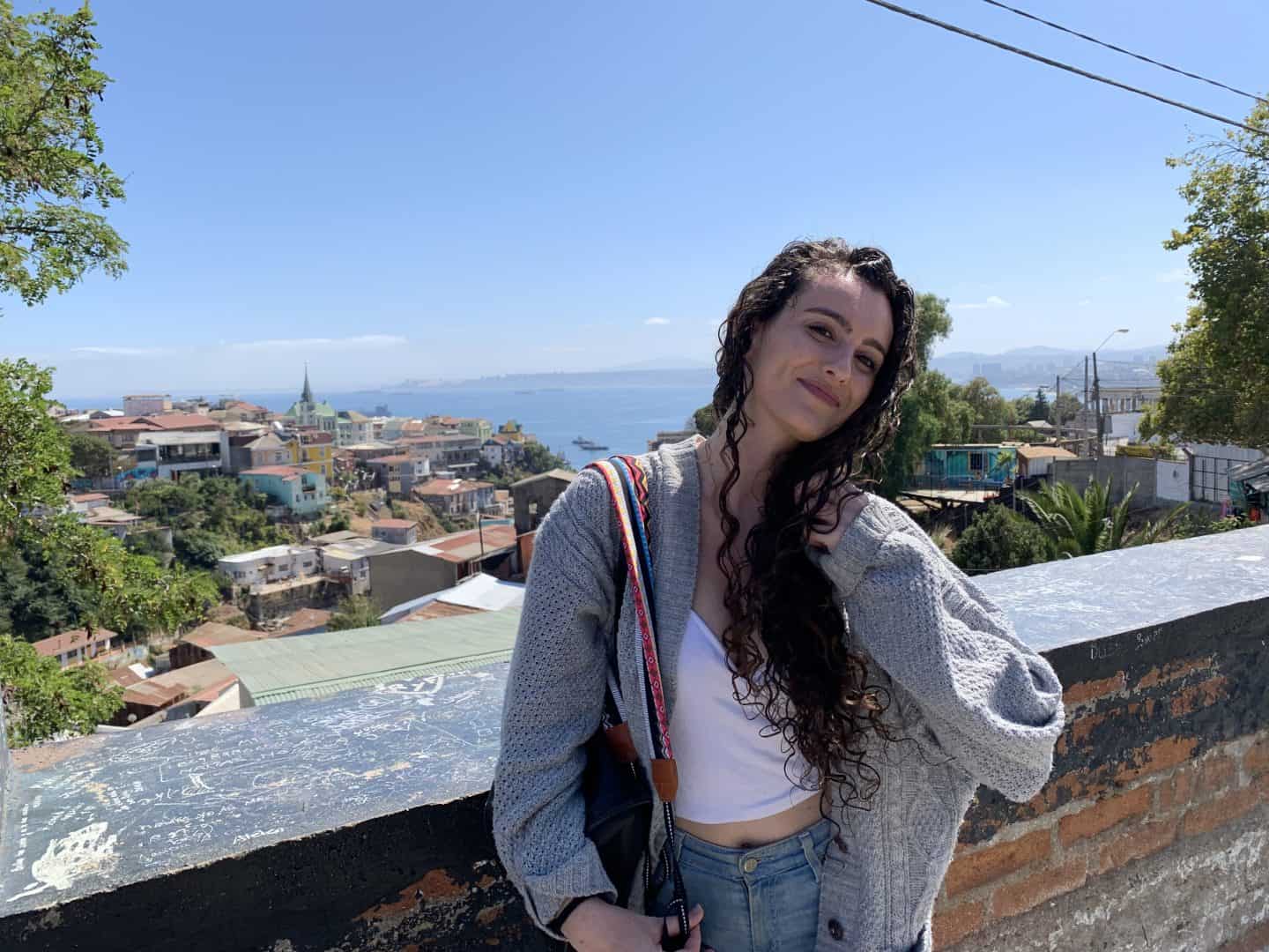 A woman wearing a gray sweater stands in front of a view of the ocean and multi-colored buildings in Valparaiso, Chile