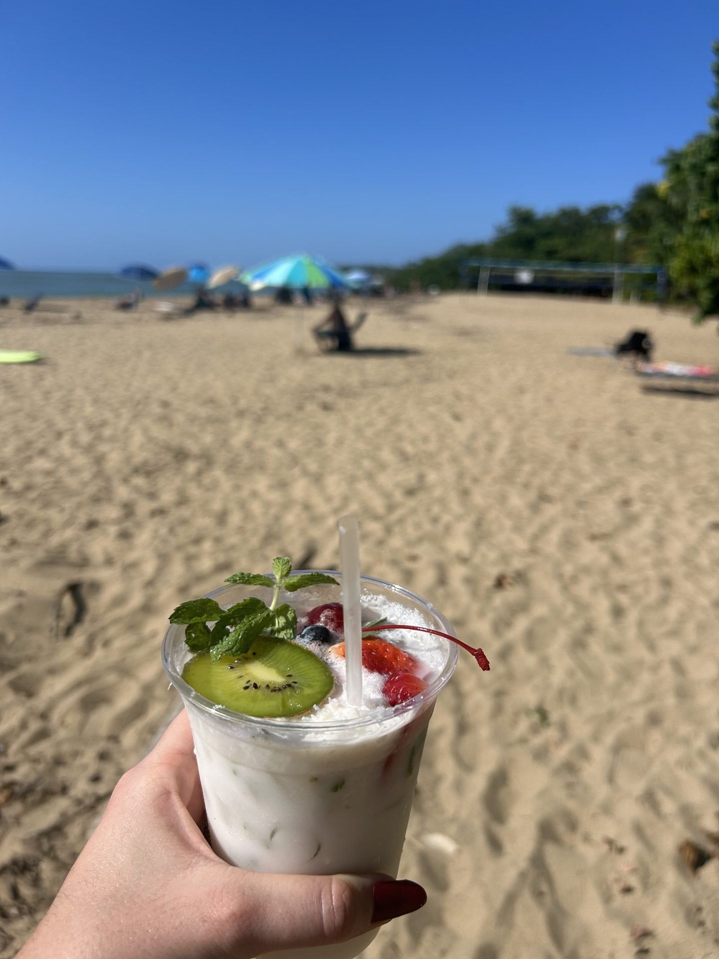 A hand with long purple nails holding a piña colada in a plastic cup. A slice of kiwi, mint, and cherries are visible at the top of the drink. A beach in Rincon, Puerto Rico, is out of focus but visible behind the drink.
