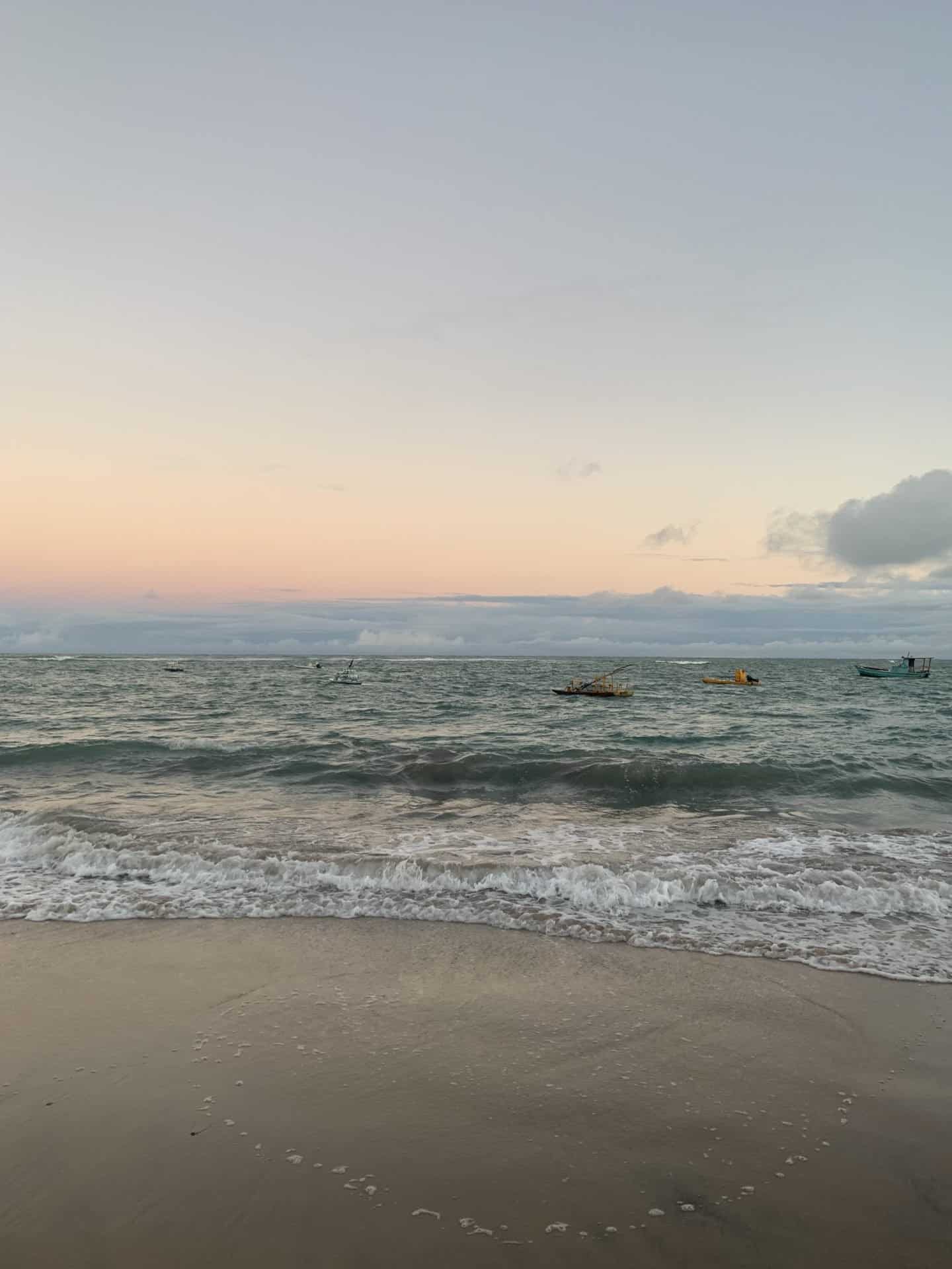 A sunset over a beach in Porto de Galinhas, Brazil, near Recife. A few boats on the ocean are visible in the distance.
