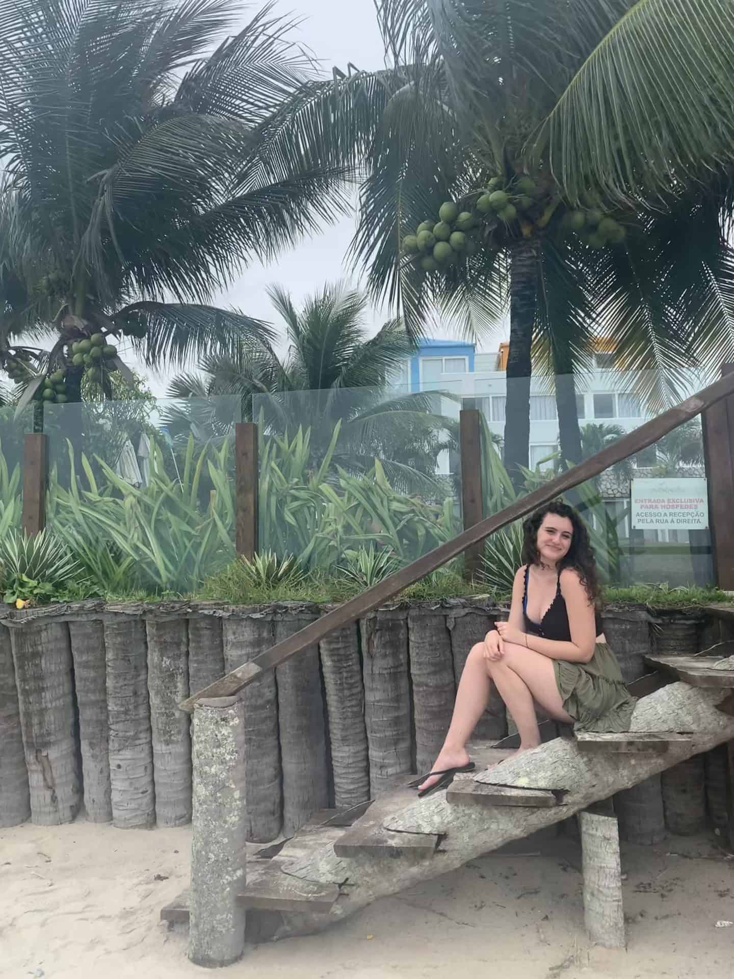 A woman with a green skirt and black shirt sitting on stairs in front of coconut palm trees in Porto de Galinhas, near Recife, Brazil.