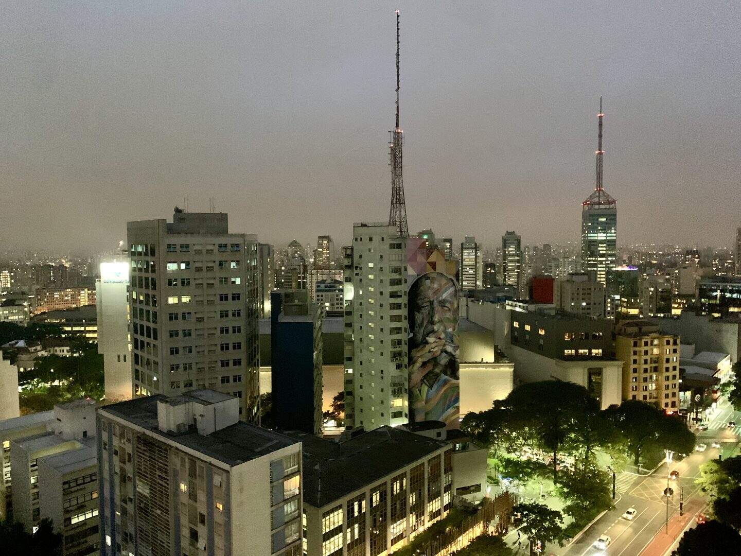 A cityscape, from SESC Avenida Paulista in São Paulo, Brazil. The sky is very gray and there are many buildings. On the building directly across the street, there is a painting of an older man's face.
