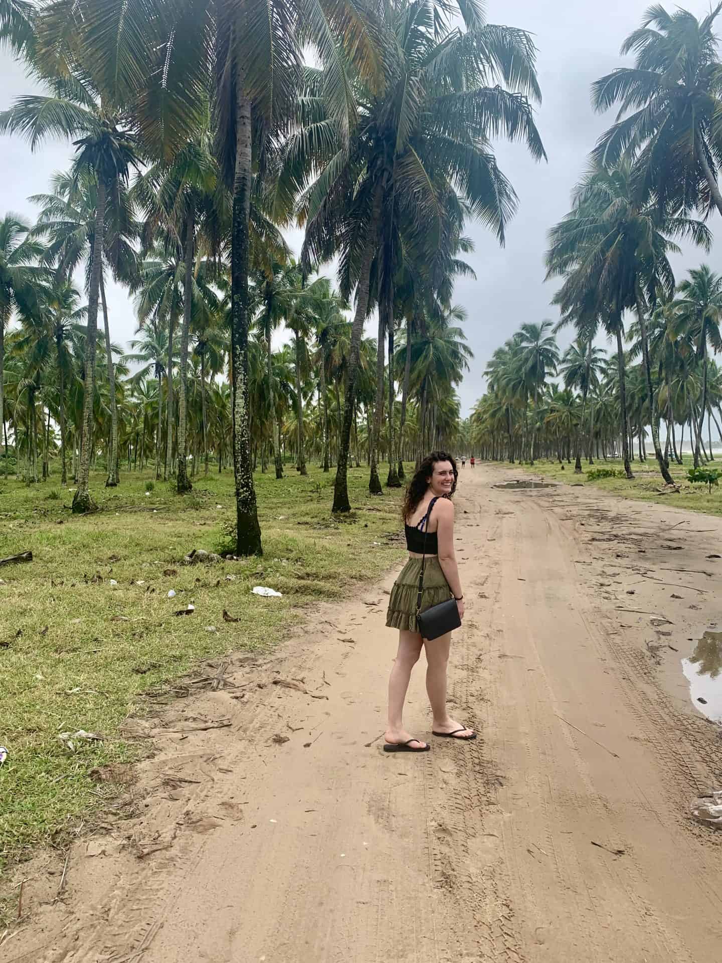 A woman in a green mini skirt and black top stands in a muddy road surrounded by palm trees.