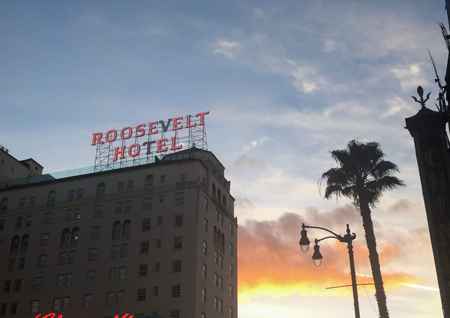 The Roosevelt Hotel at sunset, with a palm tree and a street light in shadow next to it. The sign reading "Roosevelt Hotel" it lit up in red, except for the letters "V" and the "T" in hotel, which are in black.