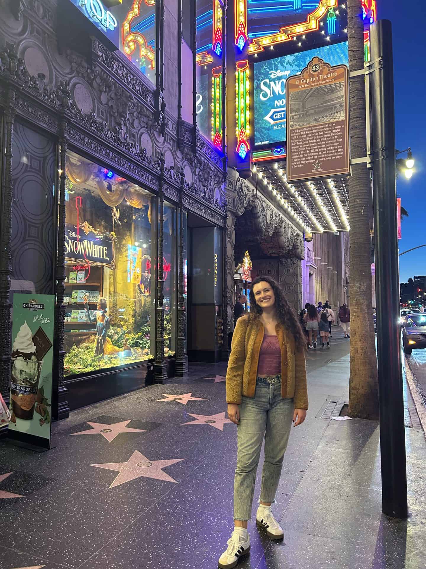 A woman wearing jeans and a brown jacket stands in front of El Capitan Theatre in Hollywood, Los Angeles, California. You can see the stars of the walk of fame, and it is nighttime.