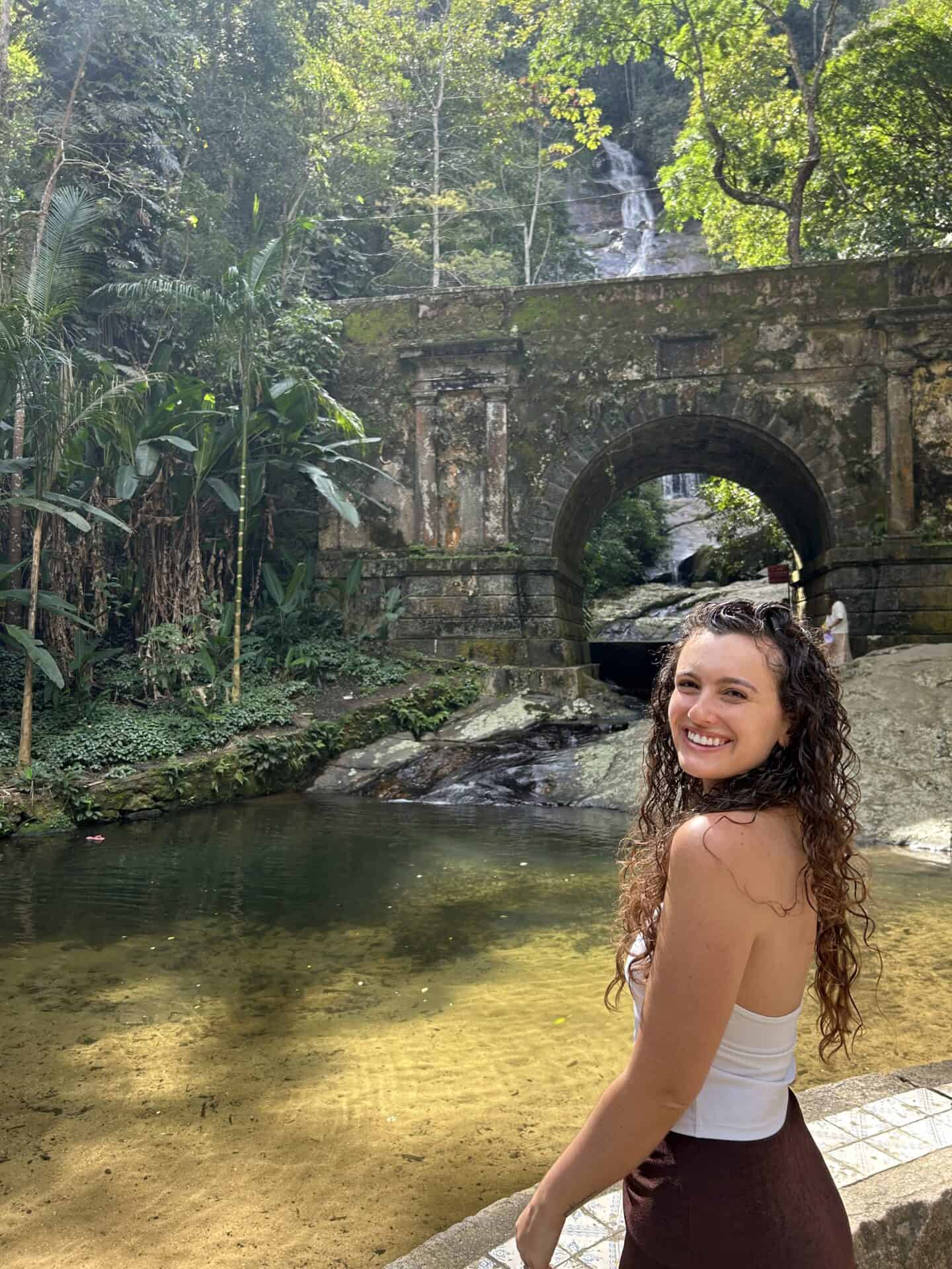 Tess, wearing a white top and a brown skirt smiles in front of a pond in Tijuca Forest, Rio de Janeiro, Brazil. Behind her are the ruins of a building with an archway, and a waterfall behind the ruins.