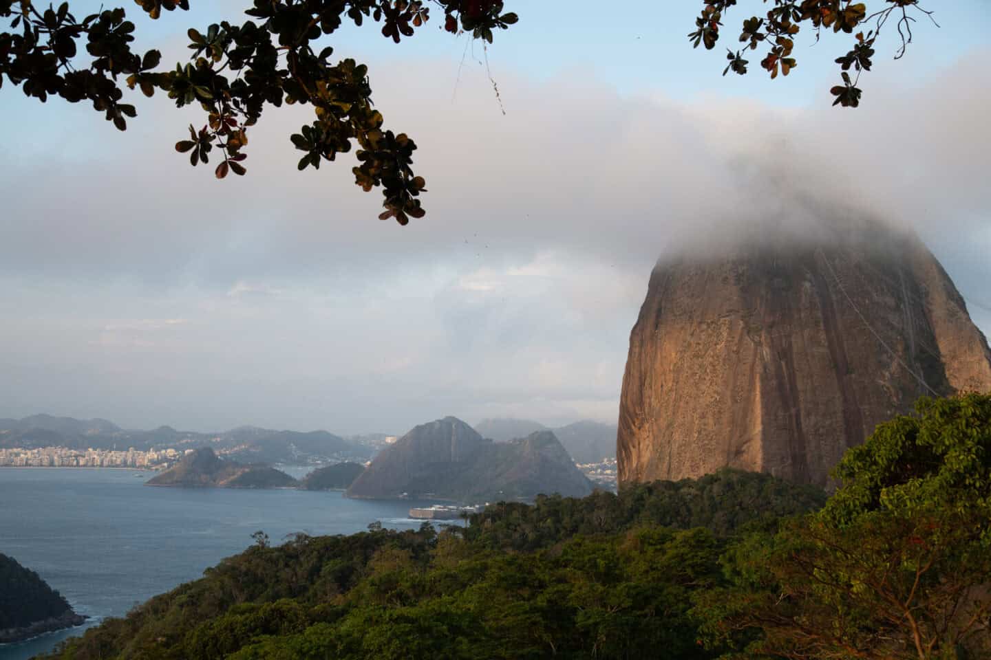 Sugarloaf Mountain with a gray cloud covering the top, with mountains, ocean, and trees below it in Rio de Janeiro, Brazil.