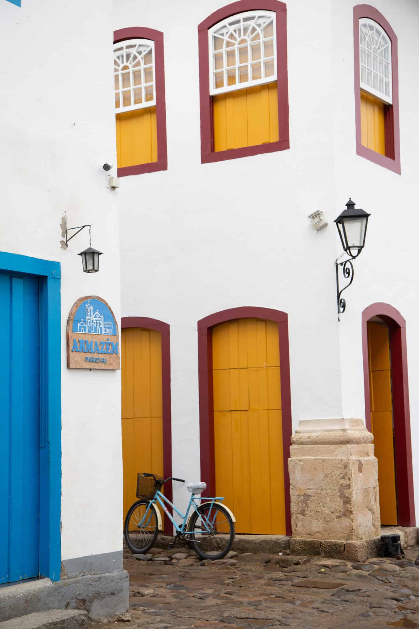 A corner of a building in Paraty, Brazil. A bike rests against the building. which has yellow doors with red outlines.