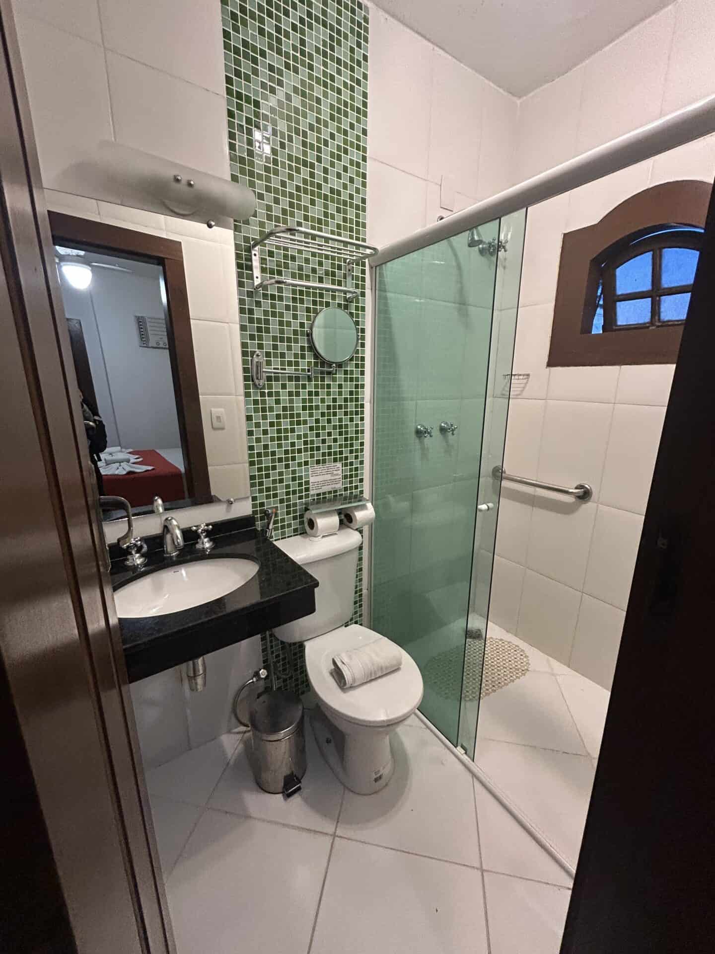 A bathroom with green tiles behind the toilet, and a shower with a green glass door, at an inn in Paraty, Brazil.