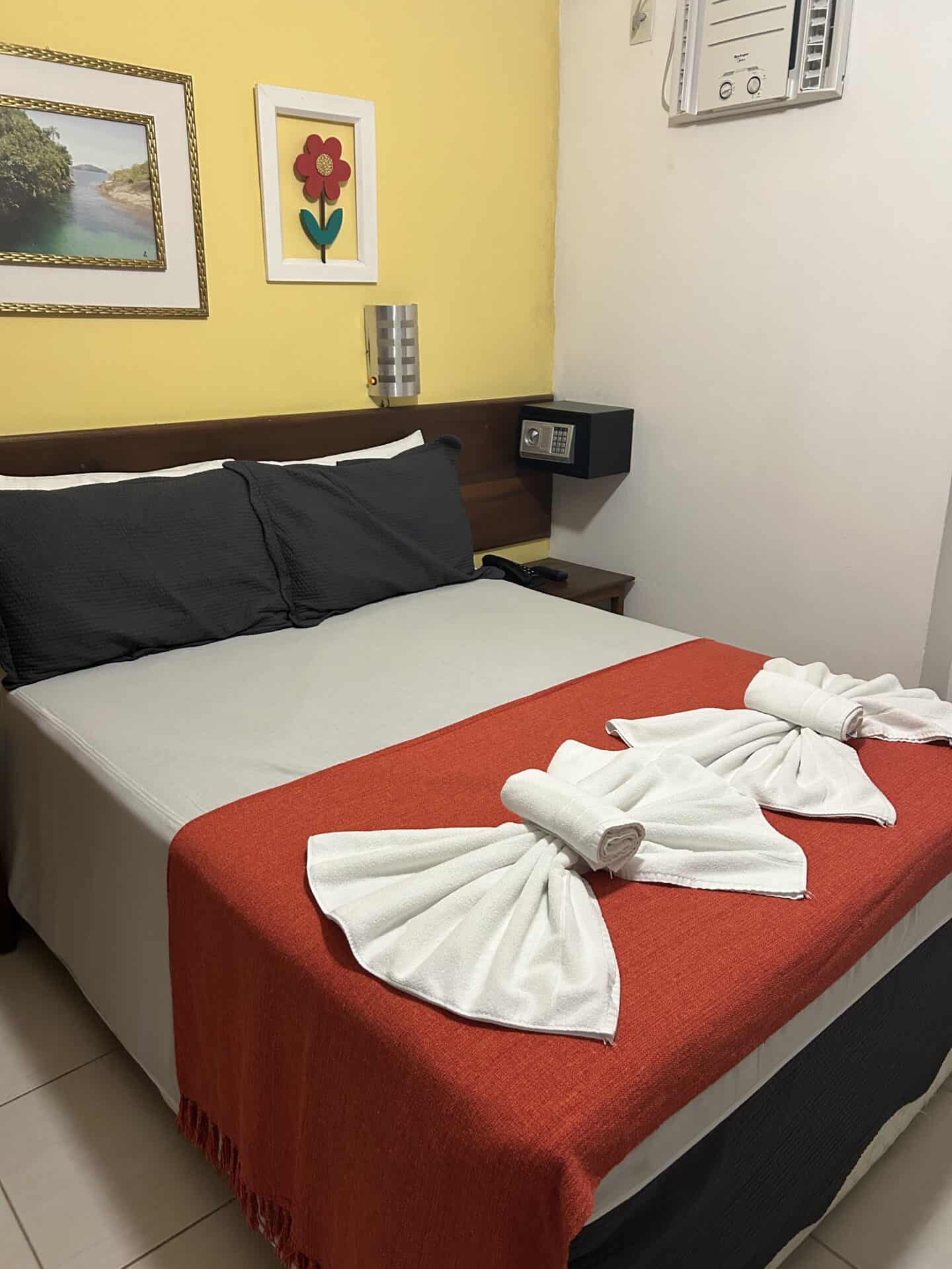 A bed with towels, and a red blanket at an inn in Paraty, Brazil. The wall behind the bed is yellow, and there is art on the wall.