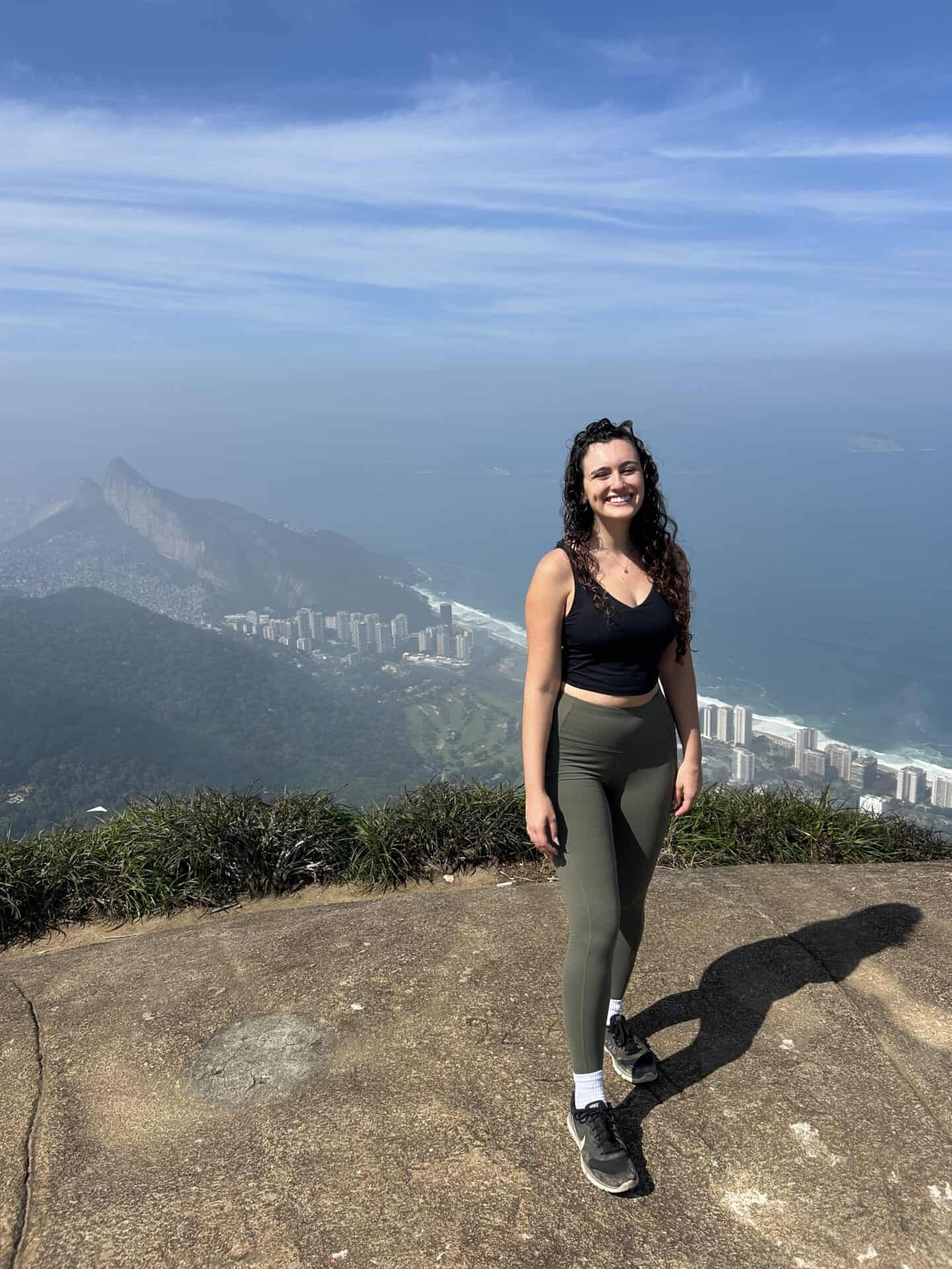 Tess, wearing green leggings and sneakers with mountains and ocean behind her in Tijuca Forest, Rio de Janeiro.