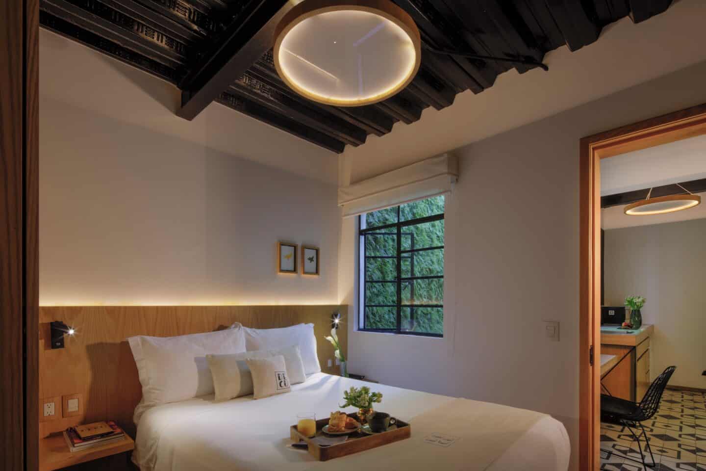 A bed with a tray on it, a window opened revealing greenery outside, and a mirror at Casa Decu in Mexico City.