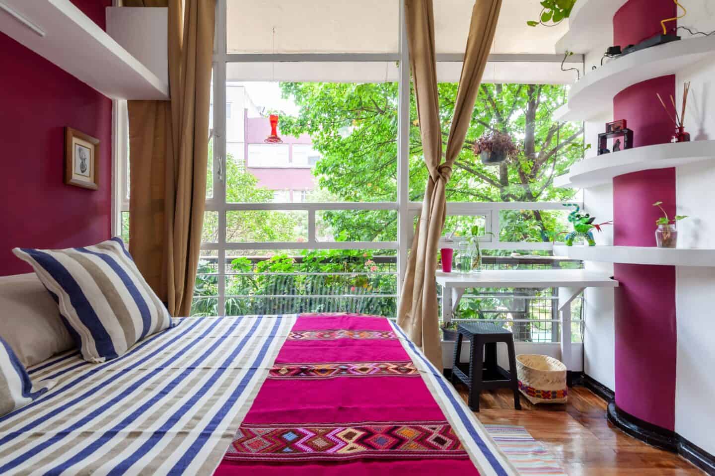 A colorful bedroom with pink and blue bedding, pink walls, and a large balcony revealing greenery at Casa Itzae B&B in Mexico City.