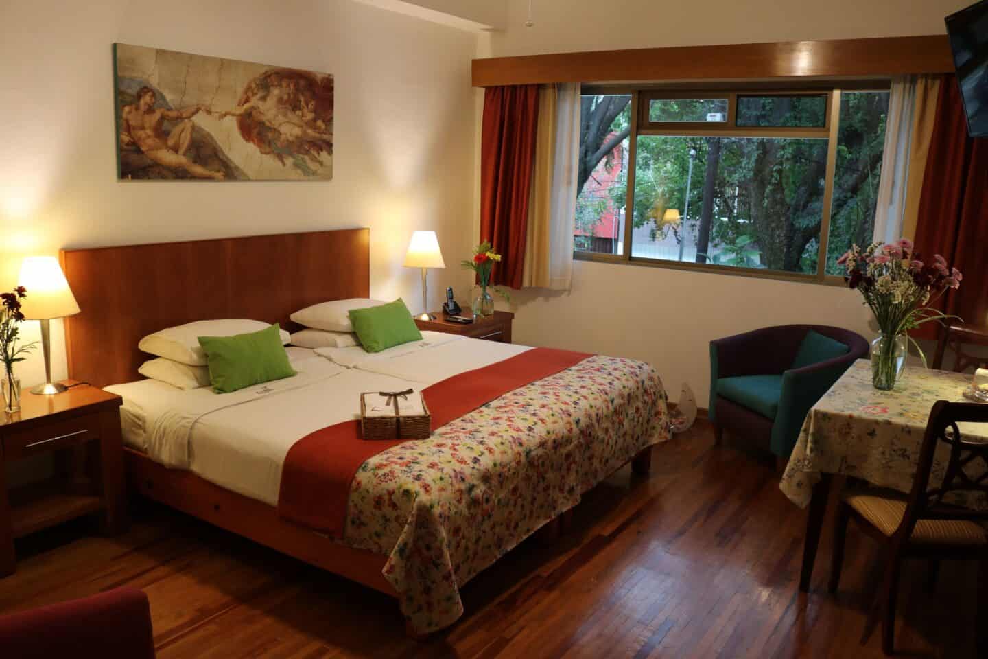 A bed with a large window, a table and chairs, and artwork at Chalet del Carmen Coyoacán in Mexico City.