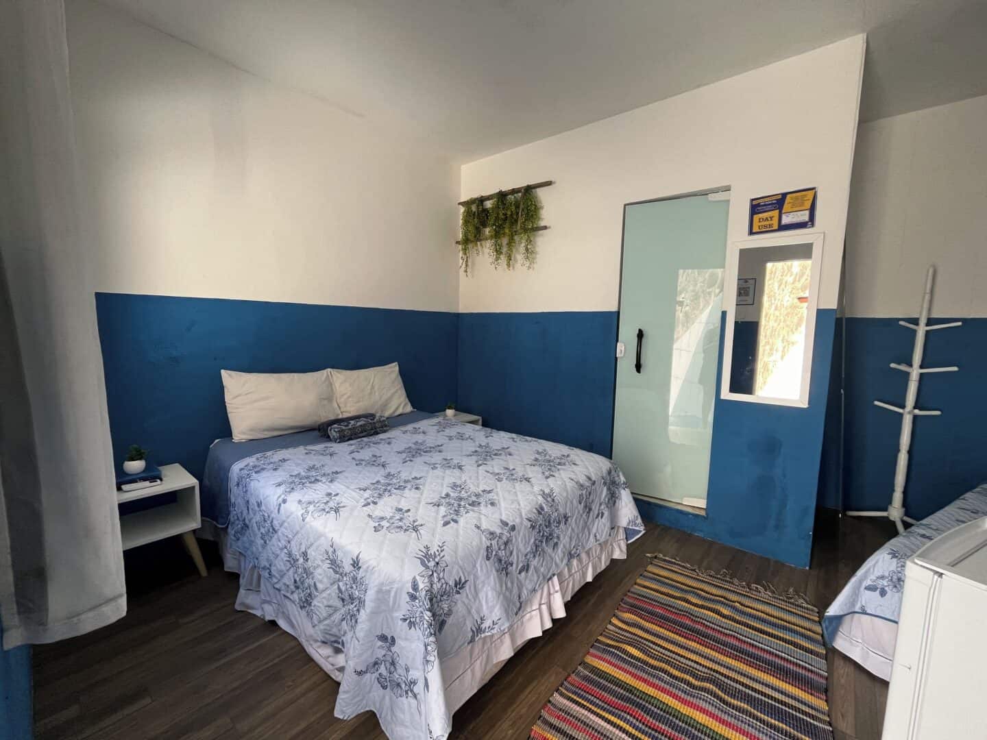 A bedroom with blue and white walls, a bed with floral bedding, a striped rug, and a bathroom door at Hostel Barra in Salvador.