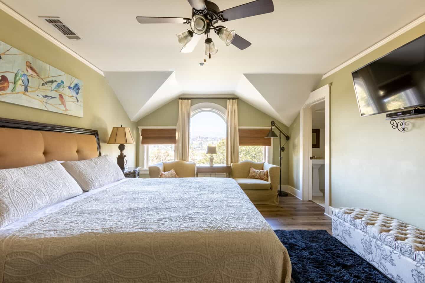A bedroom at Arroyo Vista Inn in Pasadena, California. There is a large bed with white bedding, and artwork with birds behind it. There is a television across from the bed, as well as seating, and large windows letting a lot of light in.
