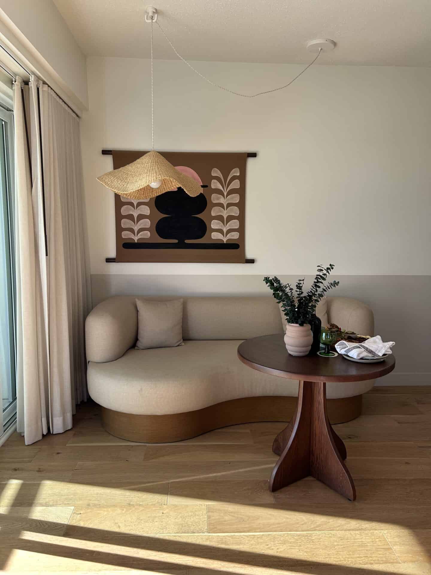 A curved couch, with a graphic design print behind it and a boho-style light, in Casa Loma Beach Hotel in Laguna Beach. A small table is in front of the couch, and light comes in from the window to the left.