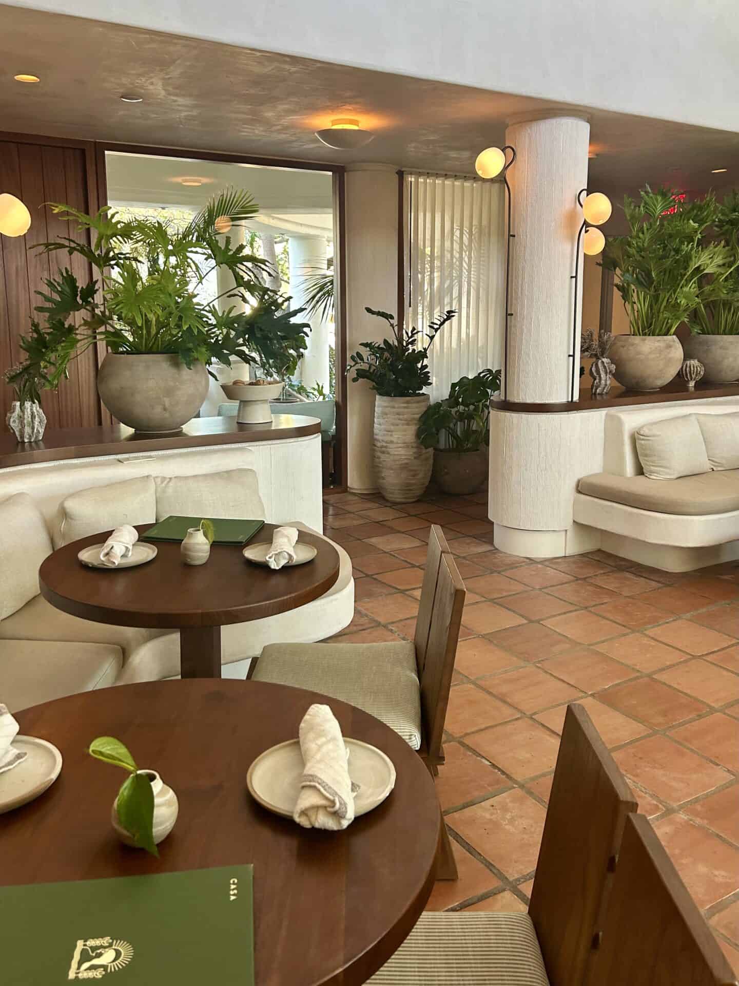 Small dining room tables with table settings in Casa Loma Beach Hotel in Laguna Beach. The room is decorated with many plants.