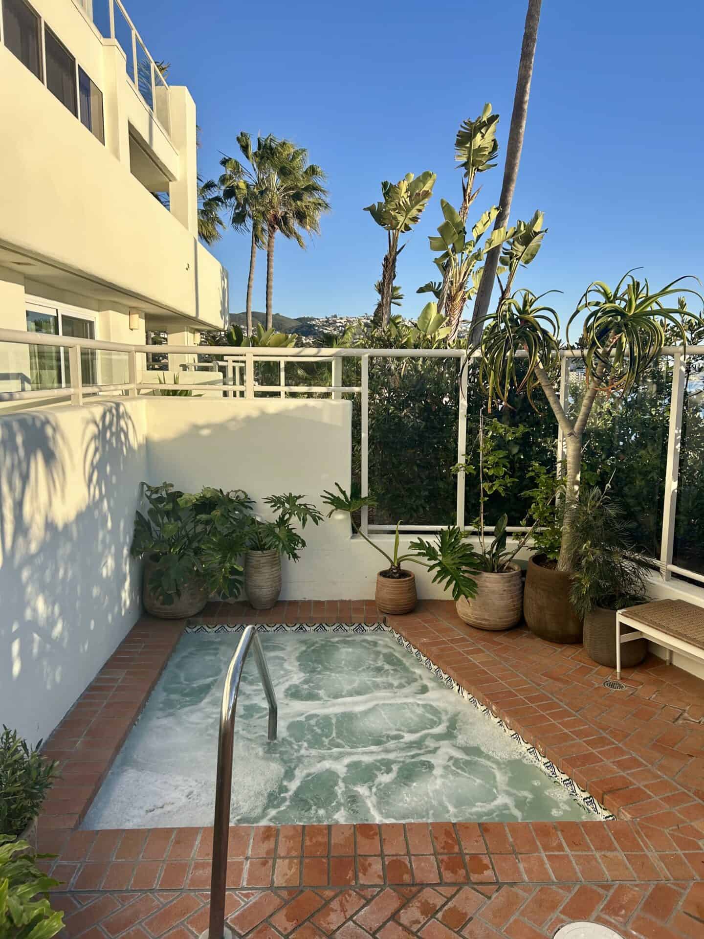 A small hot tub, in the corner of the pool area at Casa Loma Beach Hotel in Laguna Beach. Palm trees and plants are visible behind the hot tub, as well as white wall sof the hotel.