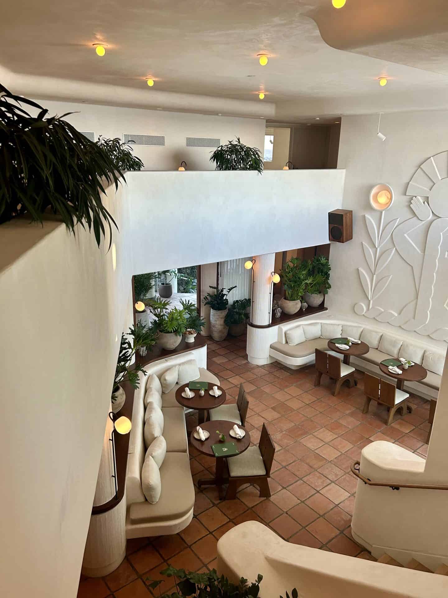 A look down at the lobby and dining room area of Casa Loma Beach Hotel in Laguna Beach. Walls are white, and a carving of a plant is slightly visible on the right side of the image. There is seating, and wooden dining tables, with many plants visible.