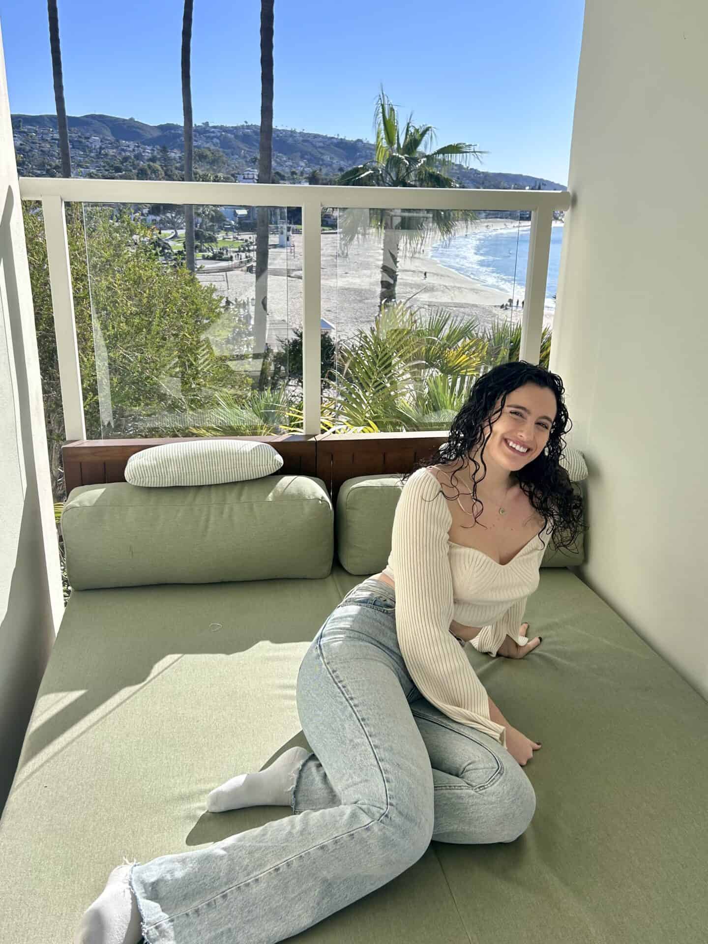 Tess, wearing a white top and jeans, sits on the balcony with the beach visible behind her, in a room at Casa Loma Beach Hotel in Laguna Beach.