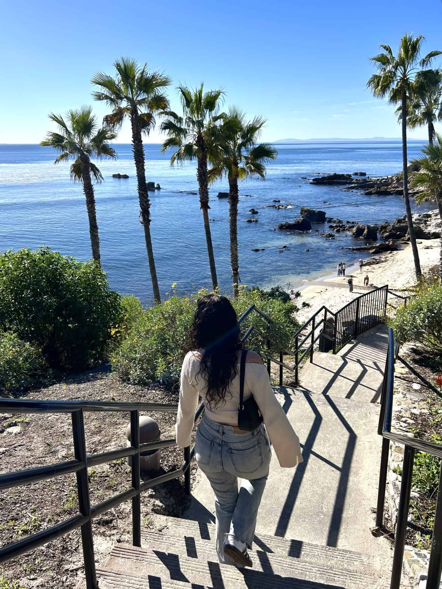 Tess, wearing a white top and jeans, walks down the stairs leading toward a rocky, palm tree-lined beach in Laguna Beach, California.