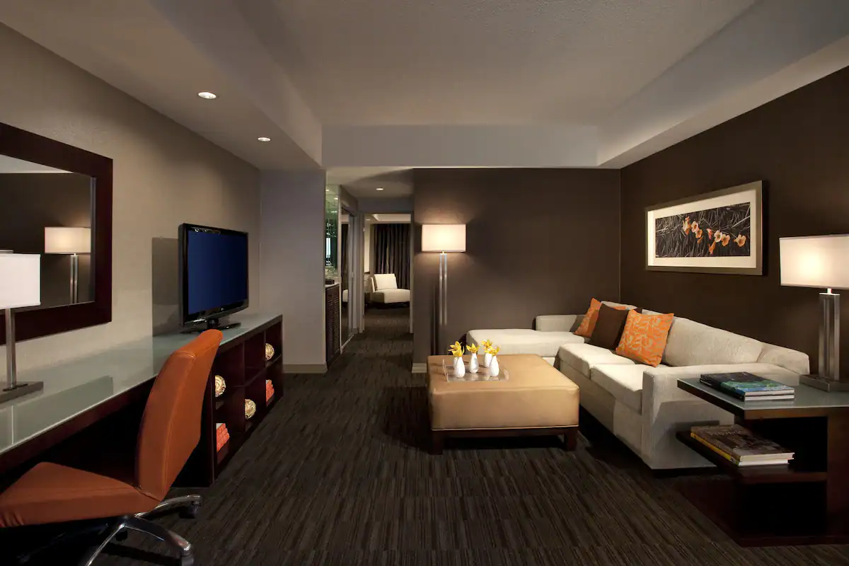A suite inside Hyatt Palm Springs. There is a couch, a desk and chair, and a television. Walls and carpet is dark.