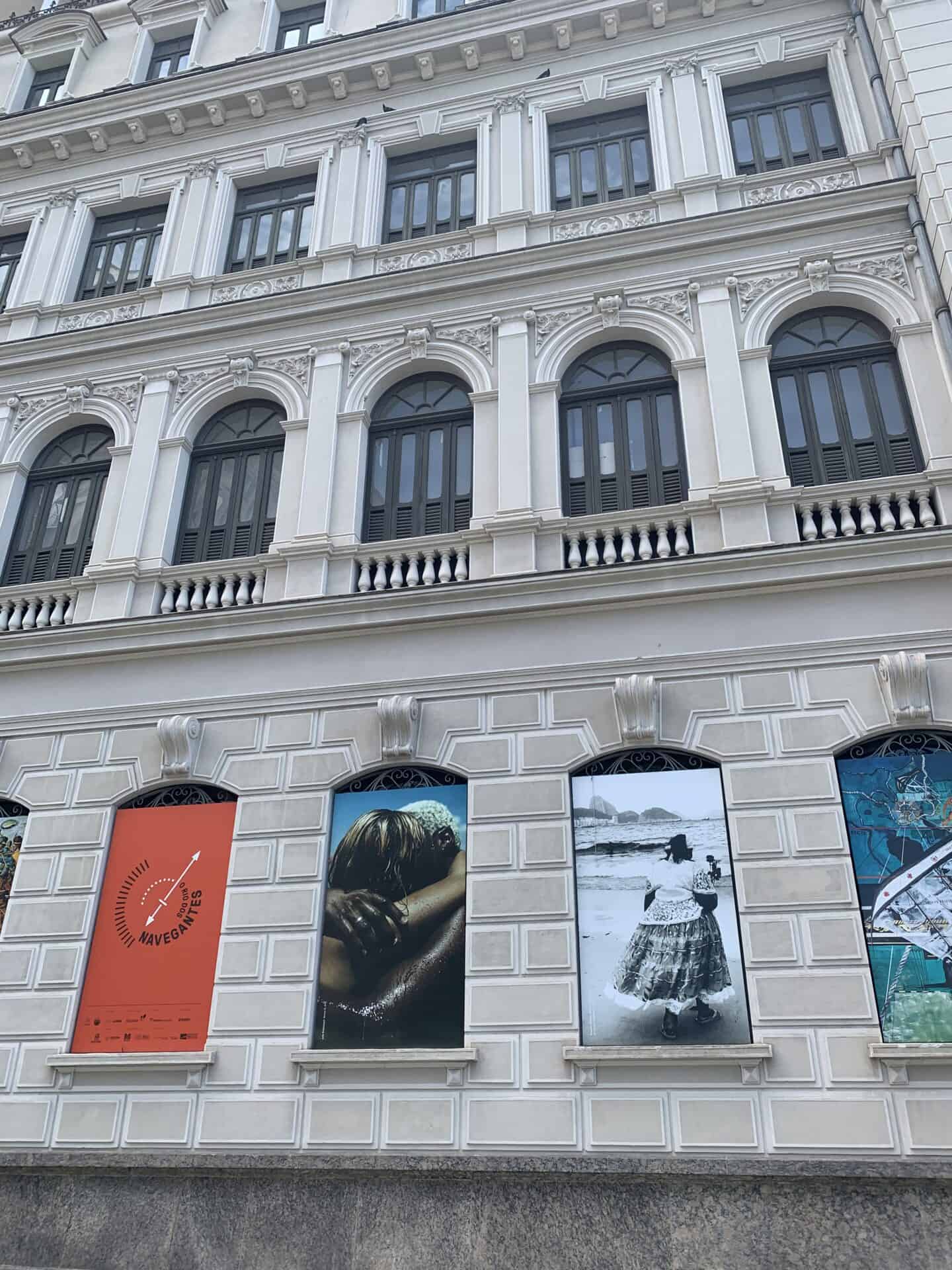 The exterior of the Rio Museum of Art in Rio de Janeiro. It is a tall, historic building made of stone. Photographs are displayed on the wall's lower level.