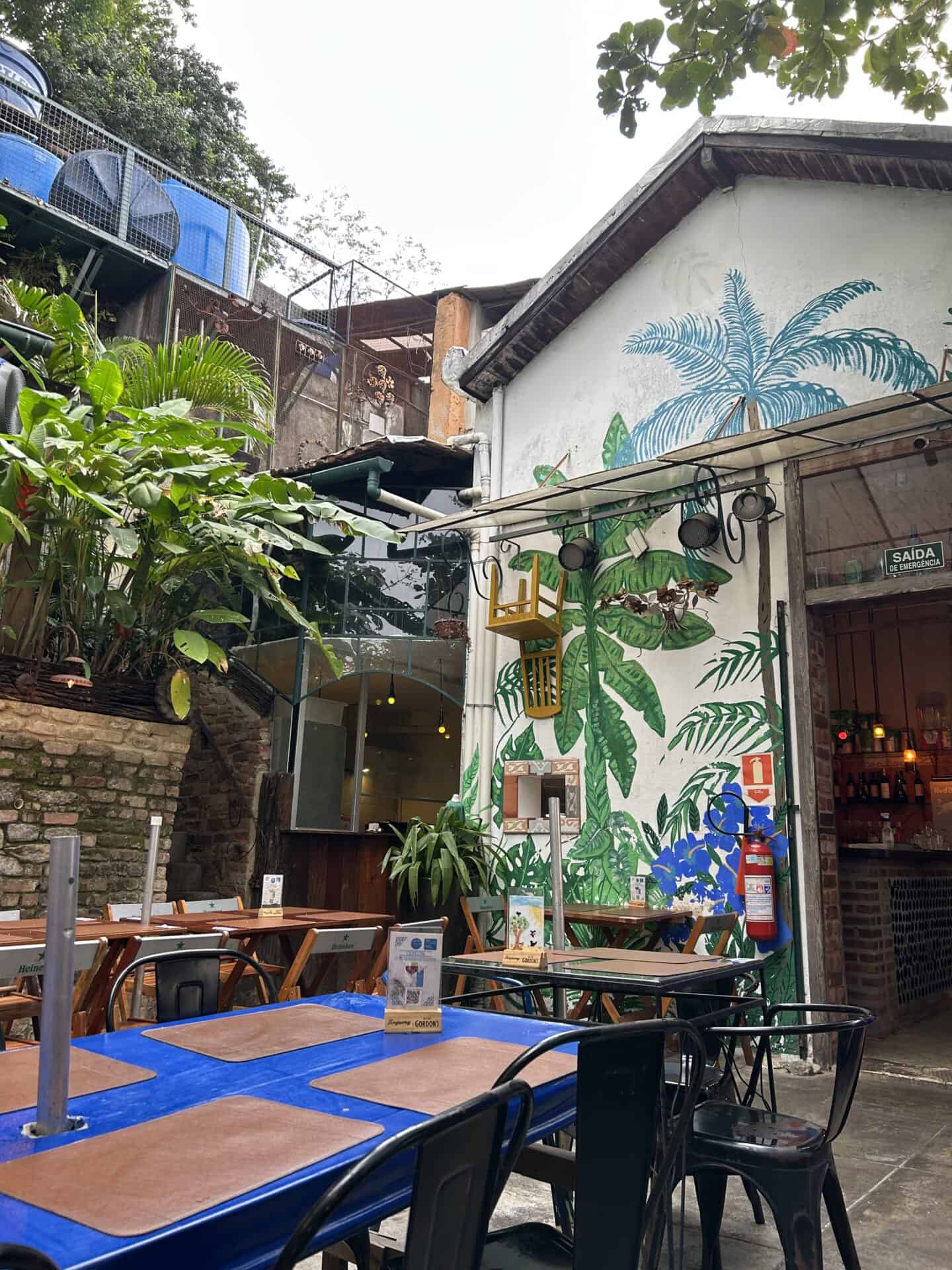Inside Casa Nossa, a restaurant in Santa Teresa, Rio de Janeiro. On the rooftop, you can see tables, many plants, and plants painted on the white wall of the restaurant.