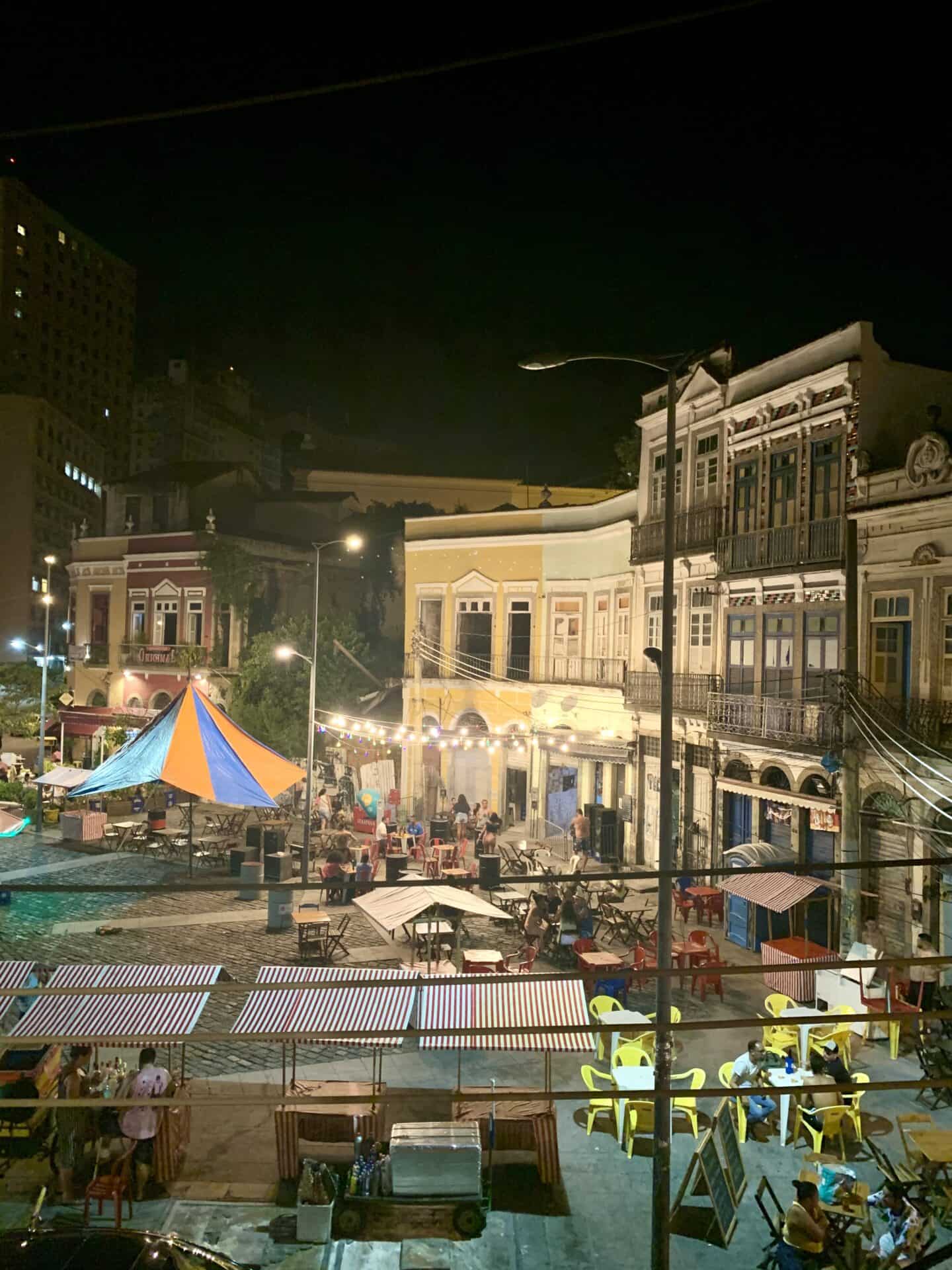 The view of Saúde, or Little Africa, from Casa Porto in Rio de Janeiro at night. The square is lit up, and many people are seated in the square, surrounded by historic buildings.